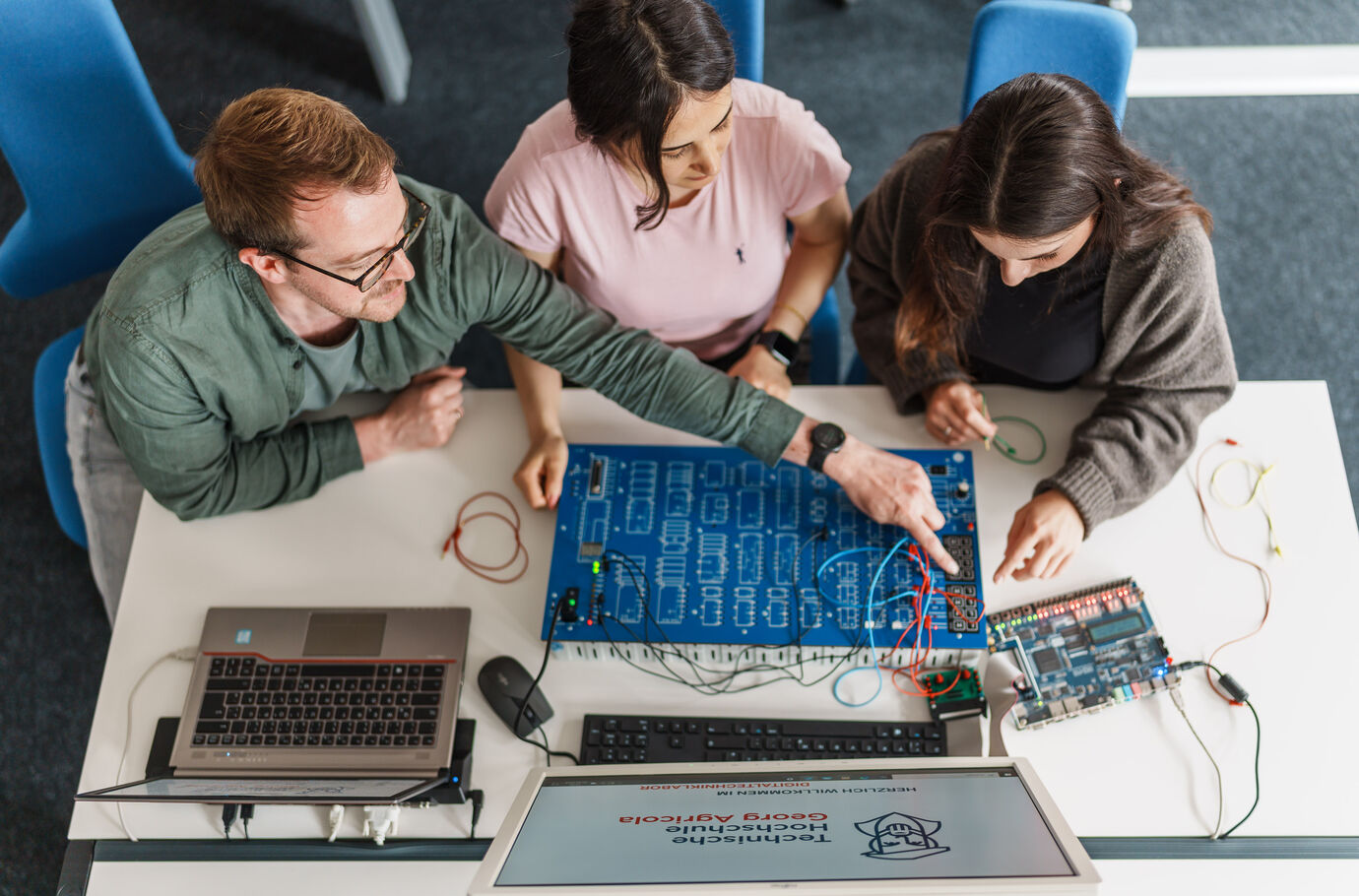 Dozent und zwei Studierende sitzen über einer elektrischen Schaltwand