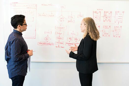 Zwei Frauen beraten vor einem Whiteboard Projektmanagementthemen für ein Unternehmen. 